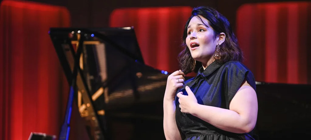 Maria Brea sings passionately on a red lit stage with a piano in the background